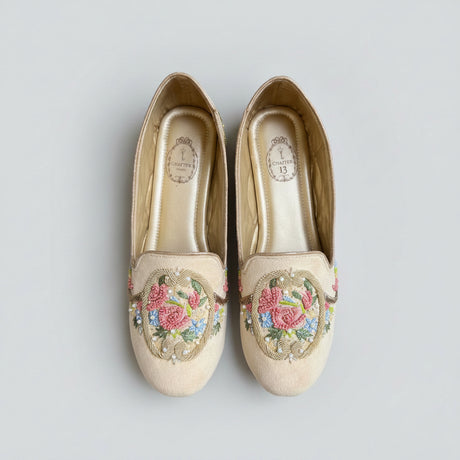 A pair of beige slip-on flats with colorful floral embroidery on the toe area, displayed on a textured gray concrete surface.