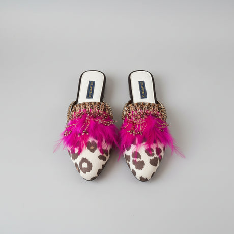 A pair of pointed-toe mule flats with white leopard-print toes, bright pink feathers, and ornate gold beaded embellishments, shown on a wooden floor. The insoles display the brand name 'CHAPTER 13'.