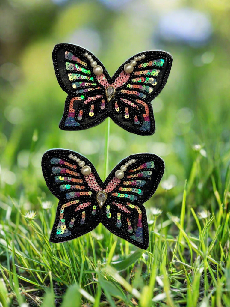 Two hand embroidered motifs of butterflies with black and multicolored wings, positioned on a grassy surface.